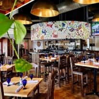 Interior of a modern Mexican restaurant with wooden tables, blue glassware, warm pendant lighting, and a colorful hand-painted mural creating a lively dining atmosphere.