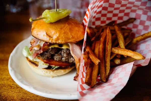 A juicy hamburger topped with bacon, lettuce, tomato, and a pepper sits on a plate next to a serving of crispy French fries in a red-checkered paper basket.