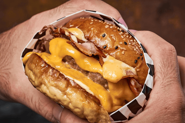 A hand holds a delicious hamburger with melted cheese, crispy bacon, and a sesame seed bun, showcasing a juicy beef patty and creamy sauce.