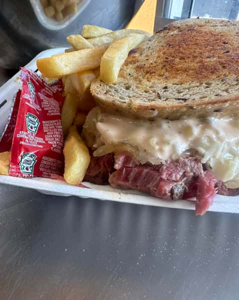 Reuben sandwich with corned beef, sauerkraut, and melted Swiss on rye bread, served with fries and ketchup packets in a takeout tray.