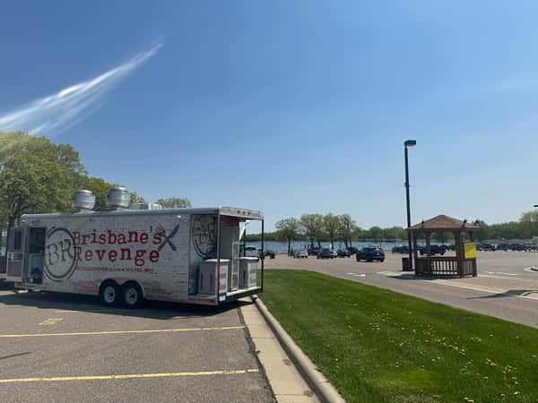 Food truck labeled "Brisbane's Revenge" parked near a lake on a sunny day in a lot with scattered vehicles and a wooden gazebo nearby.
