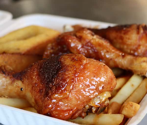 Glazed baked chicken drumsticks served over a bed of thick-cut fries in a white takeout container.