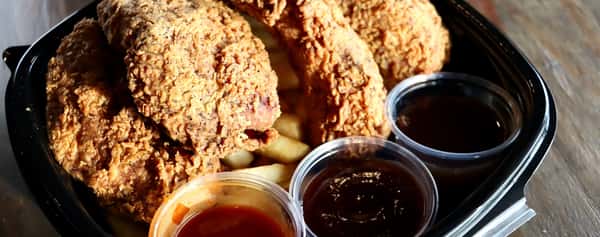 Crispy fried chicken served over French fries with three dipping sauces in a black takeout container on a wooden table.