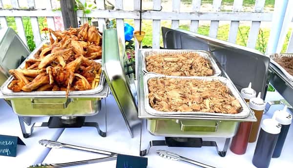 Buffet setup with trays of fried chicken wings, pulled pork, and a variety of BBQ sauces on a white tablecloth.