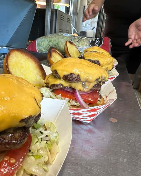That image shows several juicy cheeseburgers in paper trays, each piled high with melted cheddar cheese, lettuce, tomato, and red onion, ready to be topped with their toasted buns. They're likely being prepared in a food truck or kitchen setting.