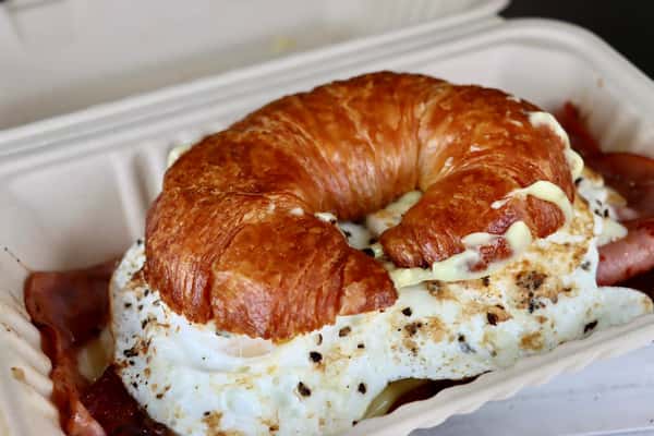 Buttery croissant sandwich filled with fried eggs, melted cheese, and ham in a takeout container.