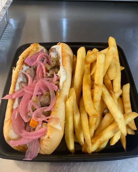 Grilled steak sandwich topped with pickled red onions and aioli, served with golden French fries in a black takeout container.