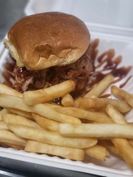 Pulled pork sandwich with barbecue sauce served alongside golden French fries in a white takeout container.