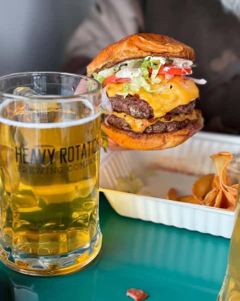 Double cheeseburger with lettuce, tomato, and onion held beside a glass of beer from Heavy Rotation Brewing Company on a green table.