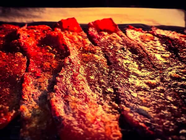 Close-up of thick-cut seasoned bacon strips glistening under warm lighting on a tray or grill.
