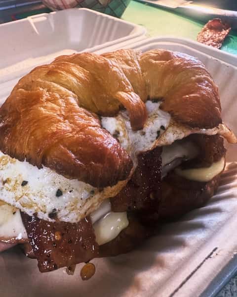 Buttery croissant breakfast sandwich filled with a fried egg, crispy bacon, and melted cheese in a brown takeout container.