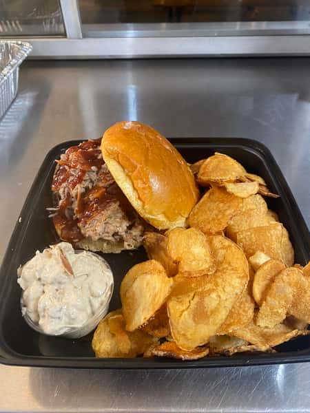 Pulled pork sandwich with barbecue sauce, kettle chips, and a side of creamy potato salad served in a black takeout container.