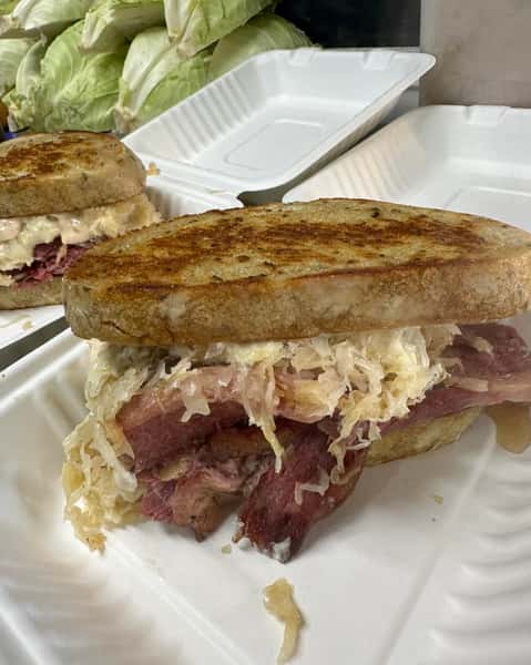 Grilled Reuben sandwich with corned beef, sauerkraut, and dressing on toasted rye bread in a white takeout container.