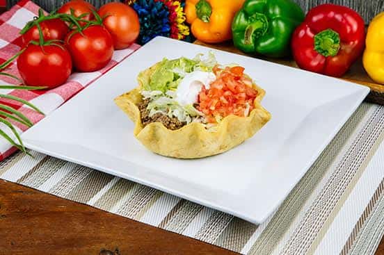 Crispy taco salad in a golden tortilla shell filled with seasoned ground beef, topped with shredded lettuce, guacamole, diced tomato, and a dollop of sour cream.