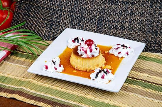 Traditional Mexican flan topped with whipped cream, chocolate and berry drizzle, and a cherry, served with extra whipped cream on a white square plate.