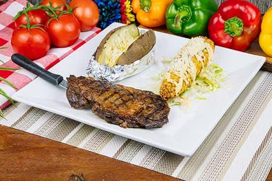 Grilled steak plate served with a baked potato wrapped in foil and Mexican-style street corn covered in cheese and spices, presented on a square white plate.