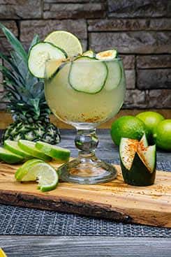 Cucumber and lime agua fresca in a goblet-style glass, garnished with sliced cucumbers, lime, and chili seasoning on the rim, presented with fresh limes and pineapple.