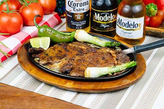 Grilled carne asada steak served on a hot skillet with grilled jalapeño, green onions, lime wedges, and accompanied by bottles of Modelo and Corona beer.