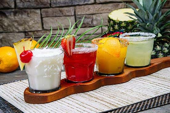 Colorful margarita flight featuring coconut, strawberry, mango, and classic lime flavors, each served in a short glass with salt or sugar rims and fresh fruit garnishes.
