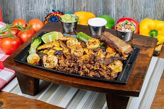 Mexican parrillada platter featuring grilled shrimp, beef, sausage, grilled onions, and nopal cactus, served with lime wedges and side dips including guacamole, sour cream, and pico de gallo.