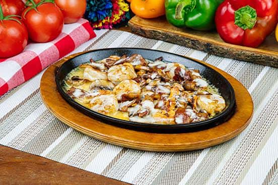 Sizzling skillet of shrimp and bacon in melted cheese, served on a wooden base and surrounded by bright bell peppers and fresh tomatoes, perfect for a hearty appetizer.