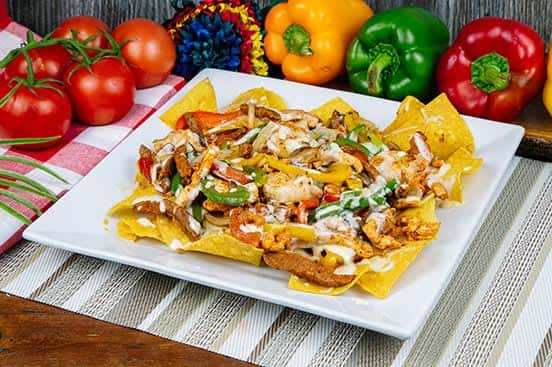 Loaded chicken fajita nachos topped with grilled peppers, onions, and drizzled with creamy white sauce, served on a white plate with a colorful backdrop of fresh produce.