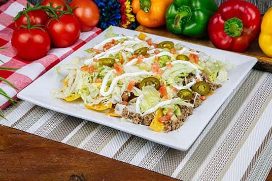 Loaded beef nachos layered with melted cheese, refried beans, shredded lettuce, tomatoes, jalapeños, and sour cream, served on a square white plate.