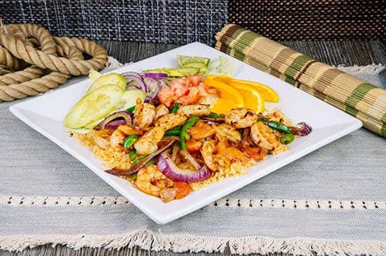 Grilled shrimp fajita platter with sautéed onions, bell peppers, and rice, accompanied by fresh cucumber, tomato, and avocado slices, served on a white plate with rustic decor.
