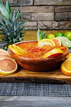 Large clay cup of Mexican fruit cocktail-style drink, rimmed with chili powder and garnished with orange, grapefruit, lime, and pineapple slices, surrounded by tropical fruits.