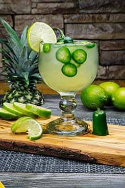 Spicy jalapeño margarita garnished with sliced jalapeños and lime in a salt-rimmed goblet, surrounded by fresh limes and a whole jalapeño pepper.