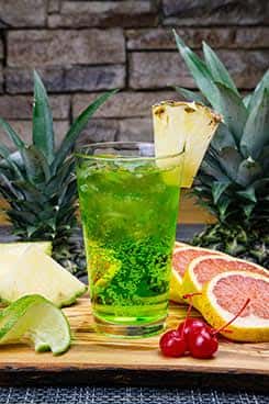 Vibrant green tropical drink garnished with pineapple wedge and citrus slices, served in a tall glass with cherries, surrounded by fresh pineapple, grapefruit, lime, and a rustic stone background.
