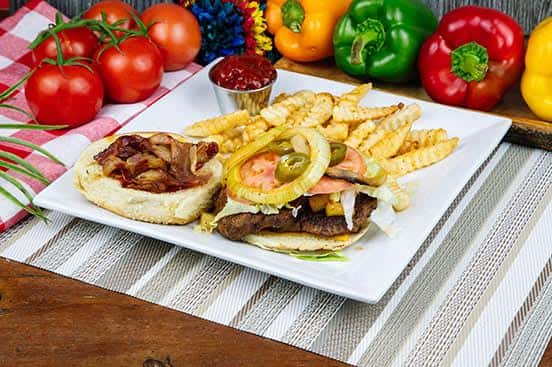 Open-faced bacon cheeseburger topped with lettuce, tomato, onions, jalapeños, and served with crinkle-cut fries and ketchup on a white square plate, surrounded by fresh bell peppers and tomatoes.
