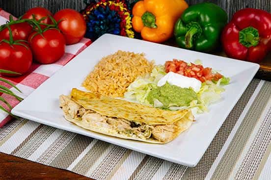 Chicken quesadilla plate with a side of Mexican rice, guacamole, sour cream, lettuce, and diced tomatoes, all served on a large square white plate.