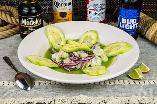 Seafood aguachile served with sliced cucumber, red onion, and avocado in a green chili marinade, presented in a white bowl with lime wedges and surrounded by bottled beers.