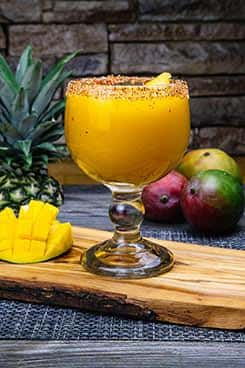 Frozen mango margarita in a chili-rimmed goblet, garnished with a mango slice and surrounded by fresh mangoes and pineapple for a tropical vibe.