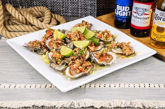 Fresh oysters on the half shell topped with a zesty seafood mix and chopped vegetables, served with lime wedges on a white square plate with Mexican beers in the background.