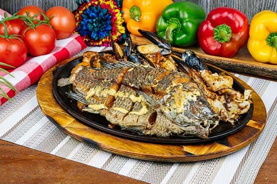 Whole fried tilapia served on a sizzling platter with grilled shrimp and mussels, surrounded by vibrant bell peppers and fresh tomatoes for a festive presentation.