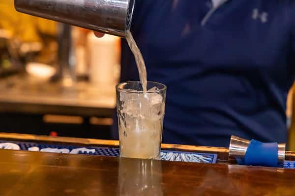 A bartender pours a freshly shaken cocktail over ice into a tall glass — likely a classic margarita or tequila-based drink in the making, perfect for complementing a flavorful Mexican meal.