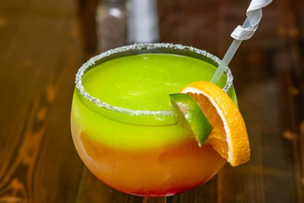 Layered frozen cocktail with vibrant green, orange, and red hues in a salt-rimmed glass, garnished with lime and orange slices.
