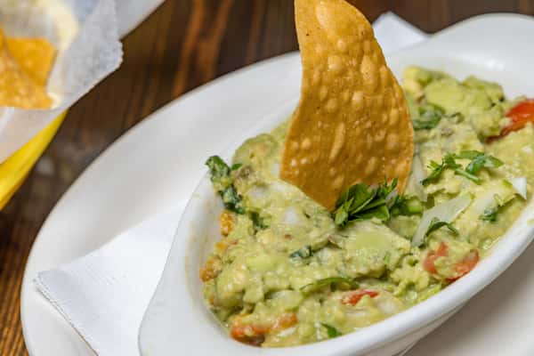 Creamy guacamole with diced tomatoes and cilantro, served in a white dish with a tortilla chip dipped inside.