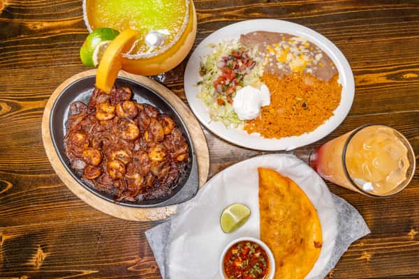 Mexican combo plate with shrimp fajitas, rice, beans, empanada, pico de gallo, and two cocktails on a wooden table.