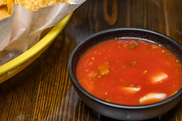 Fresh tomato salsa with onions and cilantro served in a black bowl next to a yellow basket of crispy tortilla chips.