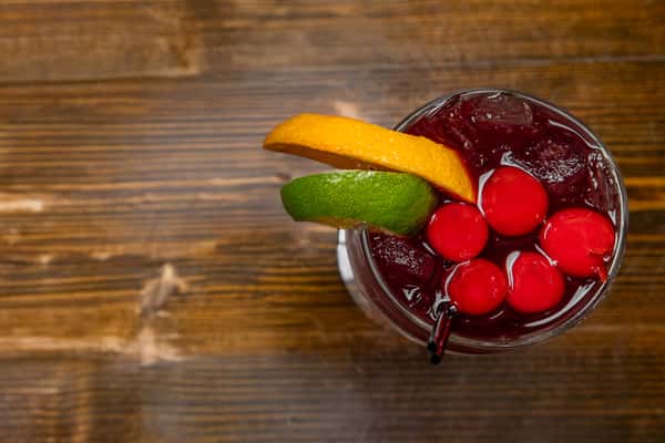 Iced cherry cocktail garnished with orange and lime slices, served in a clear glass on a rustic wooden table.