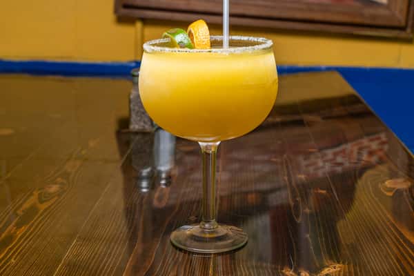 Large frozen mango margarita in a salt-rimmed glass, garnished with lime and orange slices, on a polished wooden table.