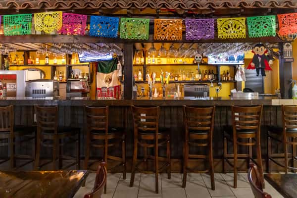 Festive Mexican bar with papel picado, wood stools, hanging glassware, and a variety of tequila bottles and beer taps on display.