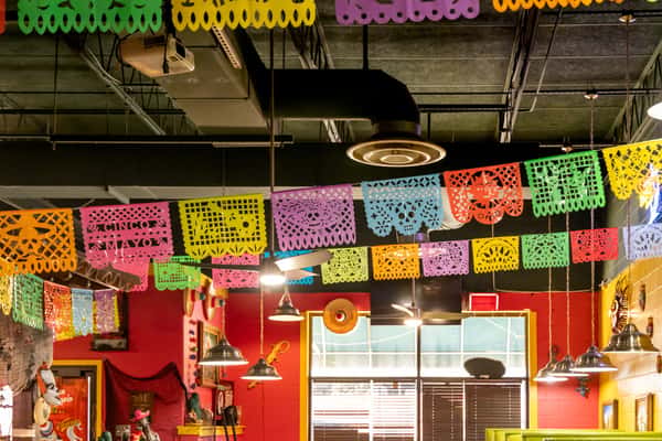 Colorful papel picado banners hang from the ceiling in a festive Mexican restaurant with red walls and industrial lighting.