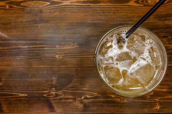 Iced cocktail with a fizzy golden hue, served with a black straw in a clear glass on a rustic wooden table.