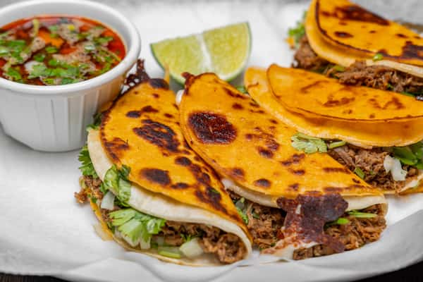Birria tacos filled with seasoned shredded meat, grilled to a golden crisp, served with cilantro, onions, lime wedges, and a rich consomé for dipping.