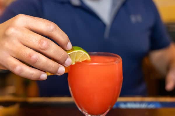 Person garnishing a bright red cocktail with lime and orange slices, seated at a bar in a casual dining setting.