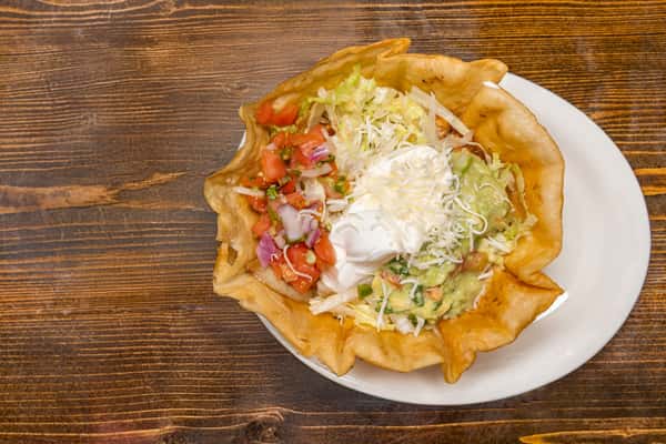 Crispy taco salad bowl filled with lettuce, guacamole, pico de gallo, shredded cheese, and sour cream on a white plate.